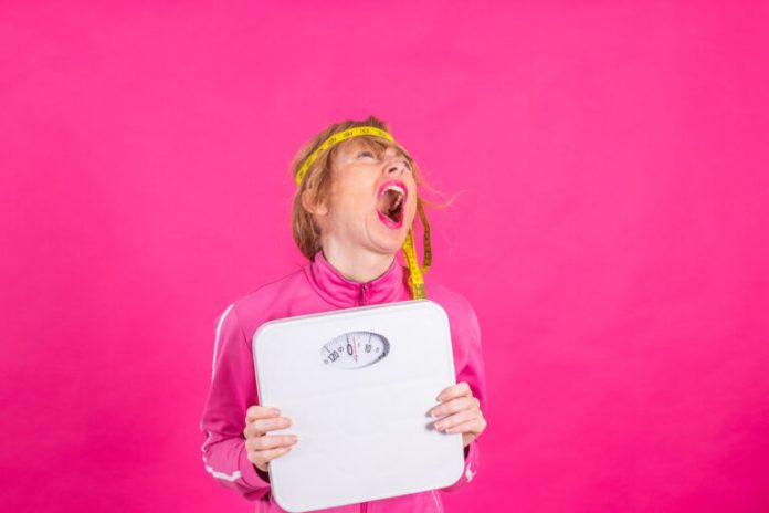 woman with weighing scale and measuring tape with expression of effort and difficulty on colored background